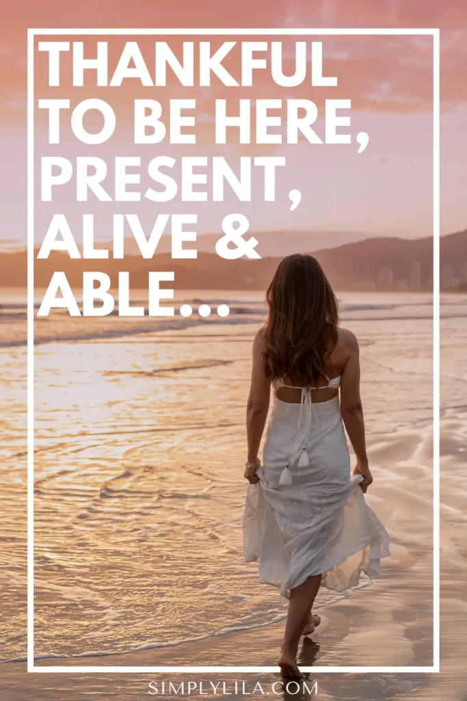 Woman walking barefoot along the shoreline at sunset, wearing a white dress, with text reading “Thankful to be here, present, alive & able,” symbolizing mindfulness, gratitude, and appreciation for life’s simple moments.